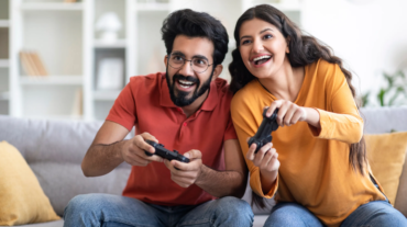 Cheerful Indian Couple Having Fun At Home, Playing Video Games Together, Happy Excited Young Eastern Spouses Holding Joysticks And Competing With Each Other While Sitting On Couch In Living Room