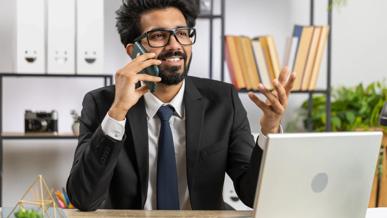 Bearded indian businessman working at office, having mobile phone talk at office desk with laptop. Manager freelance indian man holding smartphone using messenger chat apps. Employment, occupation