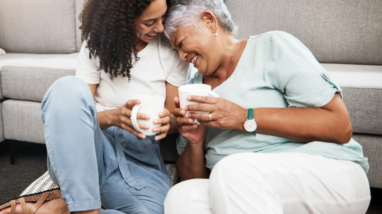 Relax, laughing and senior mother and daughter with coffee cup for home conversation, talking and bonding together. Black family, people or woman with elderly mom love, tea and living room carpet.